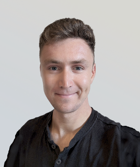 Close-up portrait of a person facing the camera against a plain light gray background. The person has short, styled hair and is wearing a dark, buttoned shirt with a soft collar. The image is evenly lit, with clear facial features and a slight closed-mouth smile. The background is clean and uncluttered, giving the image a professional headshot appearance.