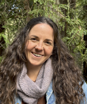 Close-up portrait of a person with long, wavy hair, standing outdoors in front of dense green foliage. The person is facing the camera with a visible smile, wearing a light denim jacket layered over a textured gray knit scarf and a warm-toned top. Natural lighting highlights the face and hair, with a soft, leafy background creating a calm, outdoor setting.