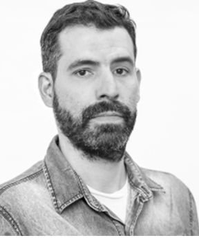 Black-and-white portrait of a person facing forward against a plain light background. The person has short hair and a full beard, and is wearing a light-colored T-shirt layered under a button-up denim shirt. The composition is a close-up showing the head and upper torso, with even lighting and minimal shadows.
