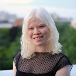 Portrait of a faculty member with shoulder-length light hair, wearing a black textured top, seated outdoors with greenery and a softly blurred campus landscape in the background.